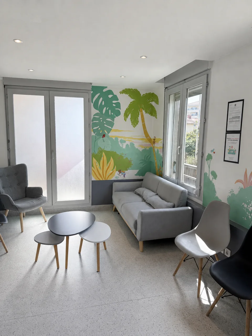 La salle d’attente du cabinet, avec un canapé gris, des sièges, de petites tables et un décor végétal peint.